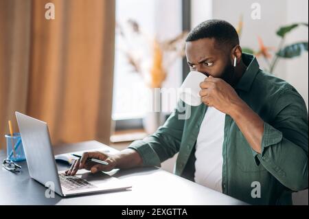 Concentrated serious busy successful African American ceo, programmer or freelancer working using laptop while sitting at his work desk and drinking a cup of coffee Stock Photo