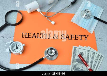 Pen, money, face mask, stethoscope, clock and magnifying glass over orange background written with HEALTH and SAFETY. Stock Photo