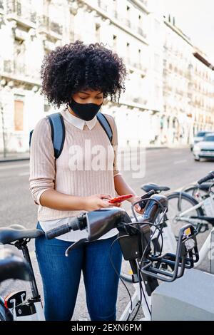 African-American woman using her smart phone to pay for a bicycle rental. Latina woman riding a bicycle in the city. Eco-friendly transportation concept. Stock Photo