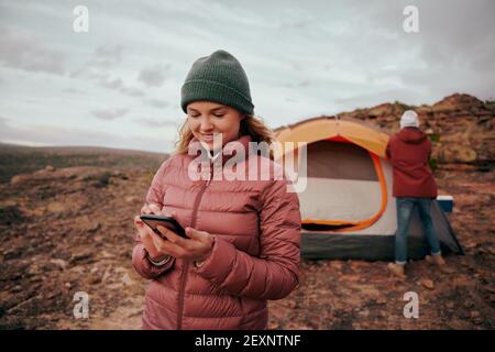 Happy young woman in winter clothing using smartphone during camping in mountain hill while man building tent in background Stock Photo