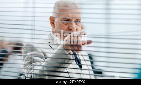 smiling Mature businessman looking through office window blinds. Stock Photo