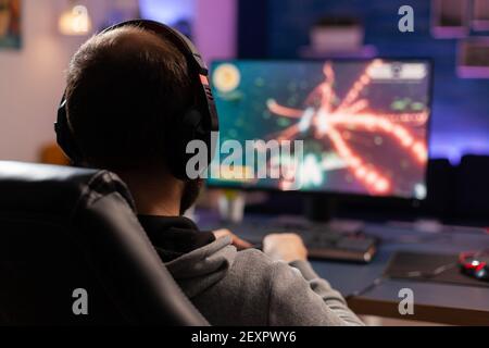 Back view shot of concentrate gamer streaming online videogames on computer using wireless controller. Player man with headphones playing games in room with neon light and professional equipment Stock Photo