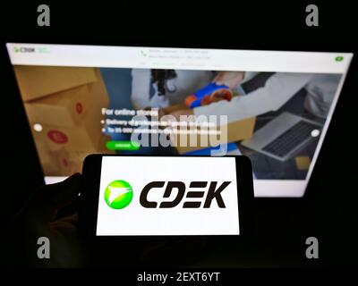 Person holding mobile phone with logo of Russian logistics company CDEK on screen in front of business web page. Focus on cellphone display. Stock Photo