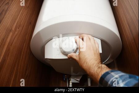 A man is switching on a domestic electric hot water boiler, water heater in a bathroom cabinet setting the temperature of the hot water for boiler eff Stock Photo
