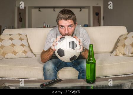 soccer supporter man enjoying game on television - nervous and attractive football fan watching tv game at home sofa couch excited drinking beer bottl Stock Photo