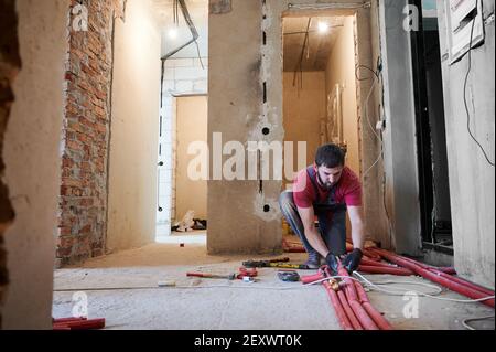 Bearded young man in gloves holding red pipe while installing heating system in empty flat with instruments on the floor. Male worker professional heating engineer working on renovation of apartment. Stock Photo