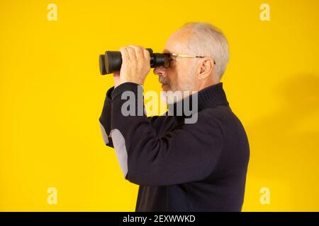 Senior man with beard and glasses using binoculars isolated over yellow background. Stock Photo