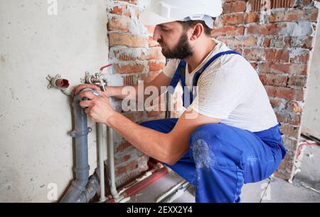 Horizontal snapshot of young plumber, wearing blue uniform and white helmet working with sealant fix of sewer pipe in kitchen or in bathroom in unfinished apartment Stock Photo