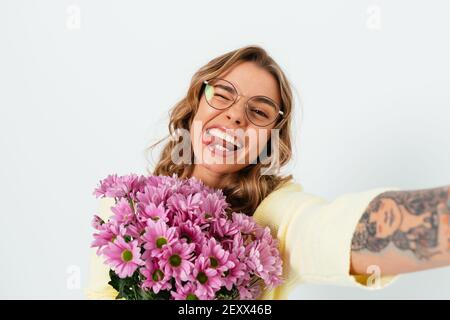 Cute young woman wearing glasses holds bouquet of fresh flowers making funny faces photographing herself standing on white background. Stock Photo