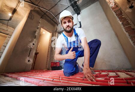 Horizontal, wide angle view snapshot of young plumber wearing blue overalls and white helmet, tying up red tubes on the floor heating system in new unfinished apartment. Giving thumb up to the camera Stock Photo