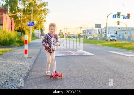 Preschooler girl riding scooter outdoors. Outdoor portrait of cute blonde hair little girl on kick scooter on a road with sunny blurred background Stock Photo