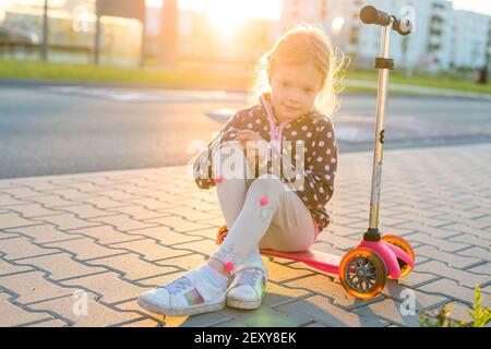 Outdoor portrait of cute blonde hair little girl sit on kick scooter on a road with sunny blurred background Stock Photo