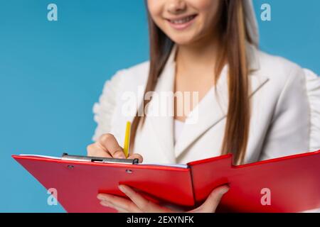 Cropped image of smiling young woman, trainer,student, business woman, consultant isolated on a blue background, writing pencil notes in work folder Stock Photo