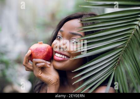 Close up portrait of young emotional beautiful afro american woman with pomegranate in her hands, looking out of green plam leaf indoors in greenhouse Stock Photo