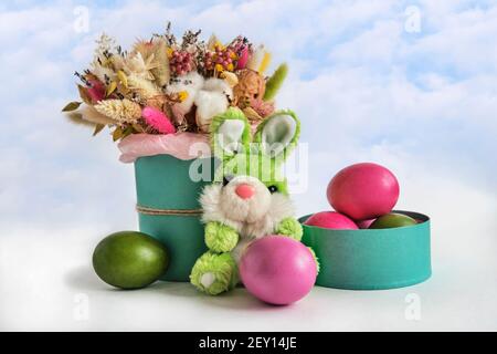 Soft toy green rabbit, beautiful bouquet of dried flowers and colorful painted natural chicken eggs. Easter composition on a cloud sky background. Stock Photo