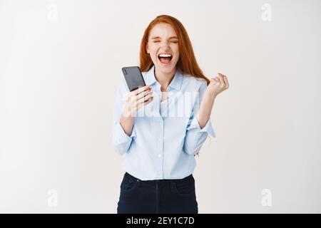 Happy redhead woman scream of joy and satisfaction, winning online prize, holding smartphone and clench one hand in triumph, white background Stock Photo