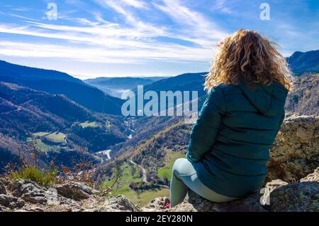 Curly-haired blonde woman, sitting on a bench, looking at the fantastic views of the Ribes mountains, in the Ripolles region, Girona Stock Photo