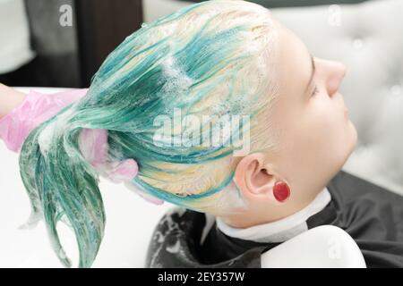 Professional beauty salon: hairstylist hands washing green hair of young woman with shampoo in sink. Head of young woman lies with closed eyes. Stock Photo