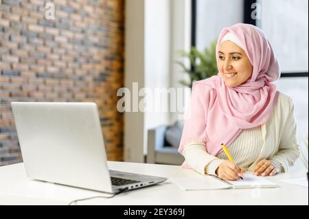 Smiling muslim businesswoman taking notes sitting at the desk in modern office space. Young islamic female student wearing hijab and smart casual wear overwrites from the laptop screen into notebook Stock Photo
