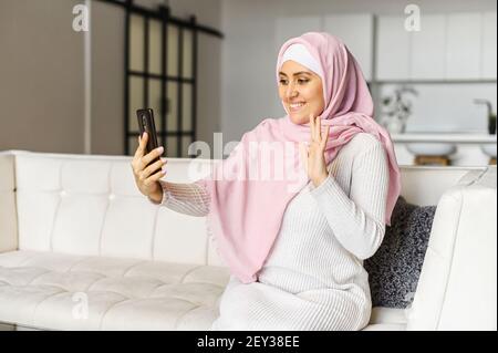 Cheerful muslim woman using smartphone for video call sitting on the comfortable sofa at home. Smiling woman wearing hijab using mobile phone for video connection with family, friends, waving hello Stock Photo