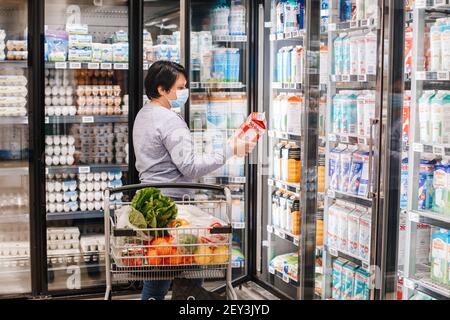 Toronto, Ontario, Canada - February 26, 2021: Grocery shopping. Middle age woman in blue protective face mask buy food milk in supermarket and put in Stock Photo