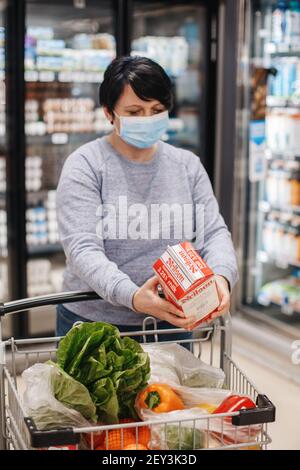 Toronto, Ontario, Canada - February 26, 2021: Grocery shopping. Middle age woman in blue protective face mask buy food milk in supermarket and put in Stock Photo