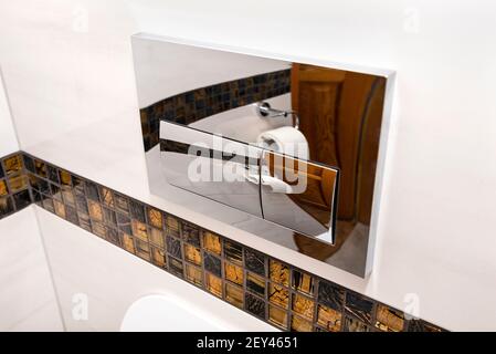 A chrome plated toilet flush with two buttons placed in the wall of a bathroom covered with ceramic tiles. Stock Photo