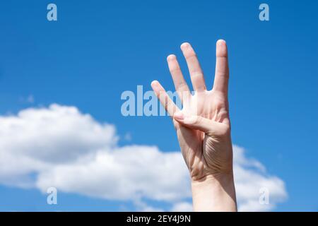 Gesture closeup of a woman's hand showing four fingers, isolated on a background of blue sky with clouds, sign language symbol number four. Stock Photo
