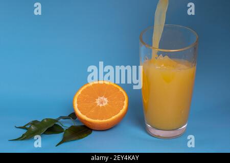 Fresh orange juice is poured into a glass, fruit cut in half and sliced in green leaves isolated on a blue background, copy space Stock Photo