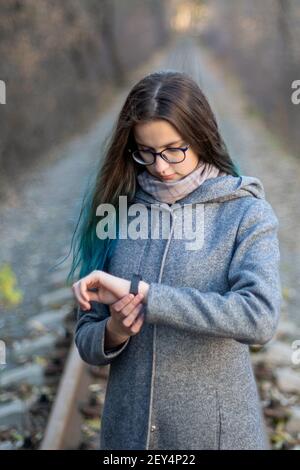 A serious European woman rushes to a meeting, looks at her watch, checks the time, dressed in a gray coat against the background of the railway in the Stock Photo