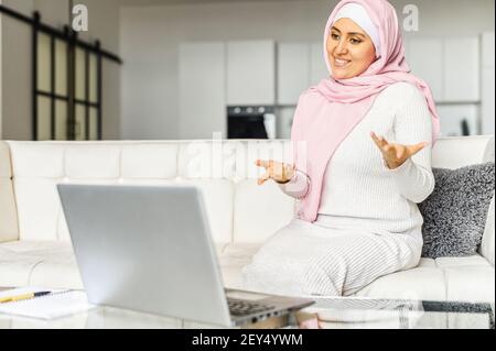 Middle-eastern woman wearing pink national hijab and smart casual wear talking online, has video chat on the laptop sitting on the couch at home, studying online. E-learning concept Stock Photo