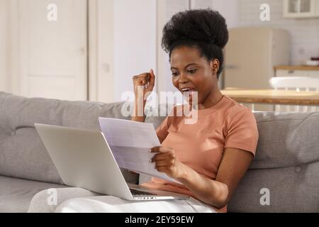 Excited Black woman sit on sofa at home receiving job enjoy exam results or college admission letter Stock Photo