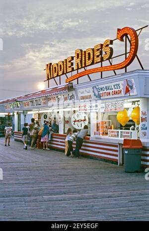 Dusk at food and ‘fun fair’ amusement stalls at Jenkinson’s Boardwalk, Point Pleasant, a borough in Ocean County, New Jersey, USA in 1974. Ice cream, cotton candy pretzels and waffles are amongst food on offer. Further along wheels of fortune offer the chance of winning toys, dolls and fish. Above a giant illuminated sign points to the ‘kiddie rides’.  This image is from an old American amateur Kodak colour transparency – a vintage 1970s photograph. Stock Photo