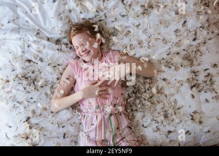 Young red haired girl laying on the bed during a feather pillow fight. Stock Photo