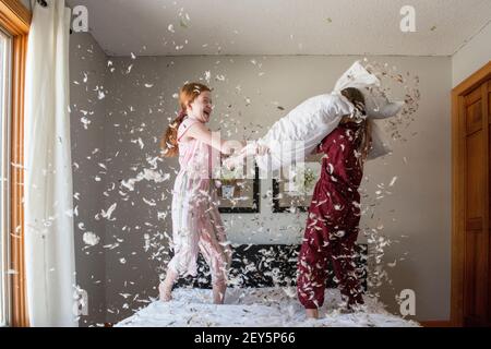 Two happy young girls having a feather pillow fight on the bed. Stock Photo