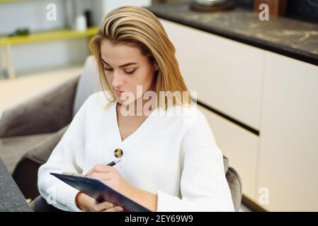 A lovely young woman is sitting at the table in her kitchen and writing in a notebook. Making a home shopping list. Stock Photo