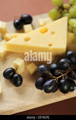 maasdamer cheese. Piece of cheese maasdam or maasdamer,green and black grapes on wooden board on brown background.Hard cheeses. Dairy products.Healthy Stock Photo