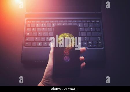 Hand hold phone with Virtual low poly hologram question mark on laptop notebook background, future technology concept Stock Photo