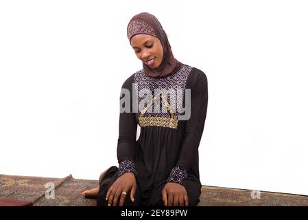 young muslim woman sitting in long dress on mat praying while smiling. Stock Photo