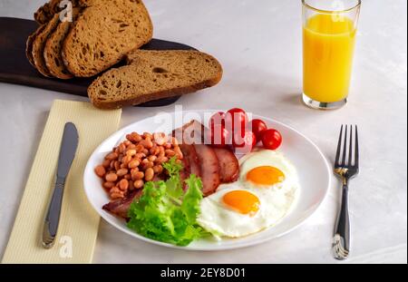 Hearty breakfast with scrambled eggs, bacon, beans, cherry tomatoes, homemade bread, orange juice on a concrete countertop. English breakfast concept. Stock Photo