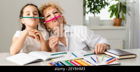 Woman tutor or foster parent mum helping cute caucasian school child girl doing homework sitting at table. Diverse nanny and kid learning writing in Stock Photo