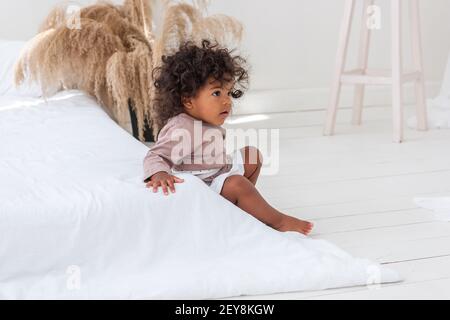 Little curly-haired African American girl plays, runs by the white bed, has fun, laughs, smiles. The concept of adopting multi-ethnic family. Scandina Stock Photo