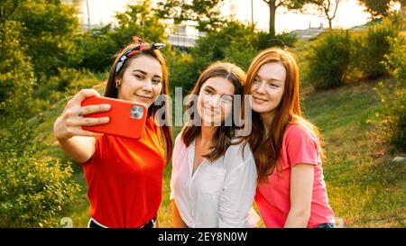 Three young girl friends taking picture of themselves on cell phone at summer sunny day. Outdoor portrait of three friends taking photos with Stock Photo