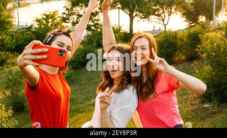 Three young girl friends taking picture of themselves on cell phone at summer sunny day. Outdoor portrait of three friends taking photos with Stock Photo