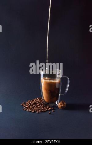 Milk pouring into black coffee in glass cup on dark blue background with beans and chocolate truffle. Closeup coffee love concept. Front view Stock Photo