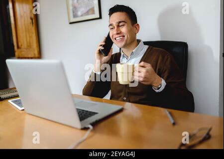 A handsome smiling young man talking on the phone holding a cup of hot coffee while looking at his laptop screen Stock Photo