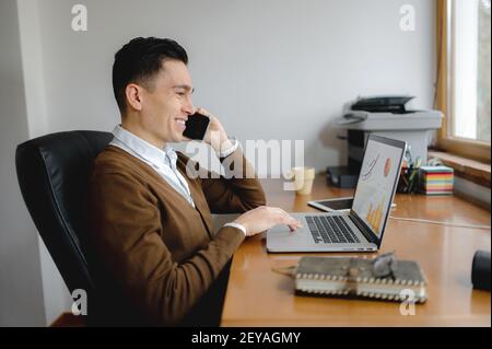 A side view of a hardworking handsome man talking on the phone with a big smile simultaneously working on his project using a laptop Stock Photo