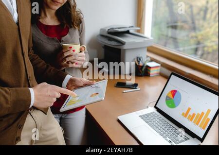 A shallow focus of two colleagues discussing business charts on a laptop and tablet in the office Stock Photo