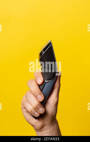 Close up on hand of unknown caucasian man holding electric beard trimmer cordless hair cutter in front of modern yellow background Stock Photo