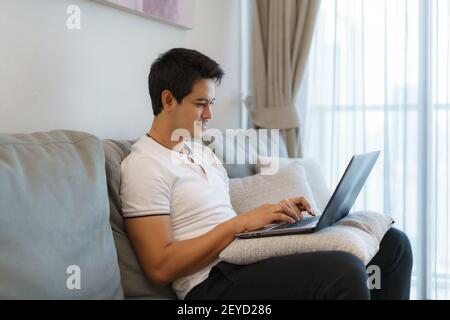 An Asian man working at home, chatting online with a colleague on his laptop while on the couch in living room at home. Stock Photo